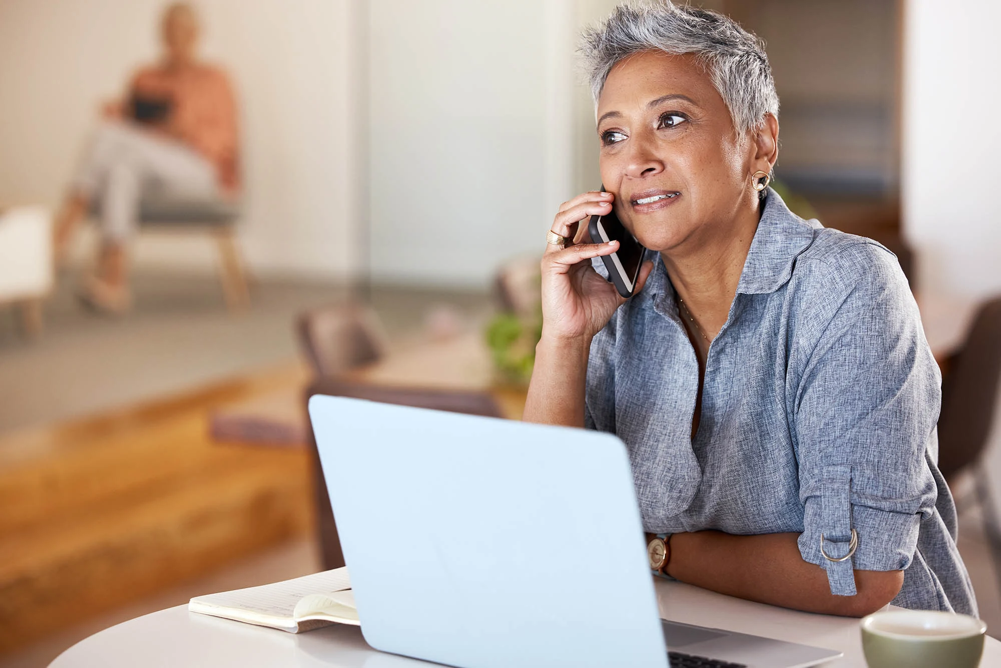 A middle-aged woman with short gray hair is smiling while speaking on her phone. She is seated at a table with a laptop in front of her and appears engaged in a conversation. A blurred figure sits in the background, focusing on their device.