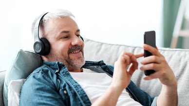 A middle-aged man with gray hair is reclining comfortably on a sofa, wearing headphones and happily using a smartphone. He appears engaged and smiling, enjoying his time with the device.