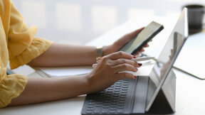 A person using a smartphone while working on a laptop at a desk with stationery and a plant in a bright environment.