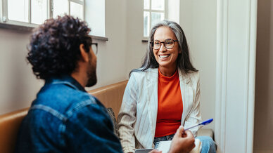 A woman with gray hair and glasses smiles while engaging in conversation with a man. She wears a red top under a white blazer. The setting appears casual, with sunlight coming through nearby windows, creating a warm atmosphere.